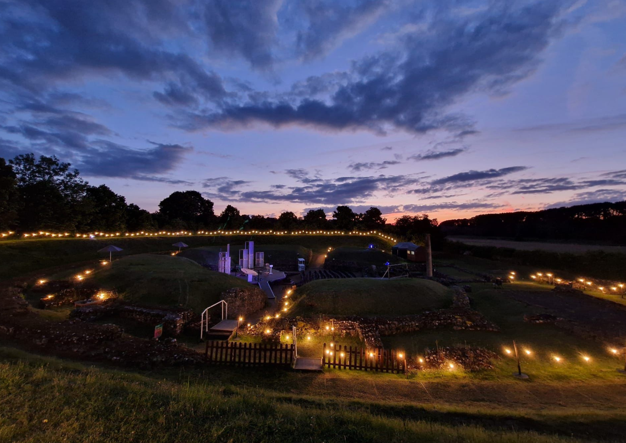 The Roman Theatre of Verulamium lit with festoon lights at dusk, its grassy earthworks and ancient stone outlines glowing beneath a wide evening sky.